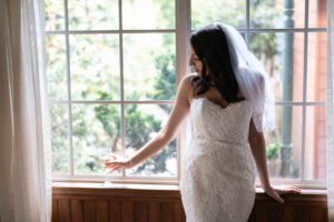 A beautiful bride with a lovely veil posing by the windowsill in the early morning, warm sunlight glowing on her