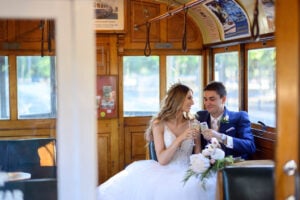 A newlywed couple toasting on a tram, with their friends cheering behind the camera